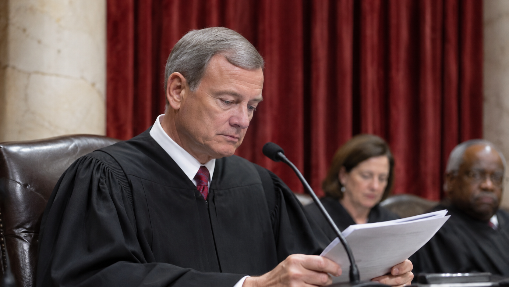 A male judge in a black robe and red tie sits at a bench, looking down at papers and speaking into a microphone, with two other judges visible in the background against red curtains.