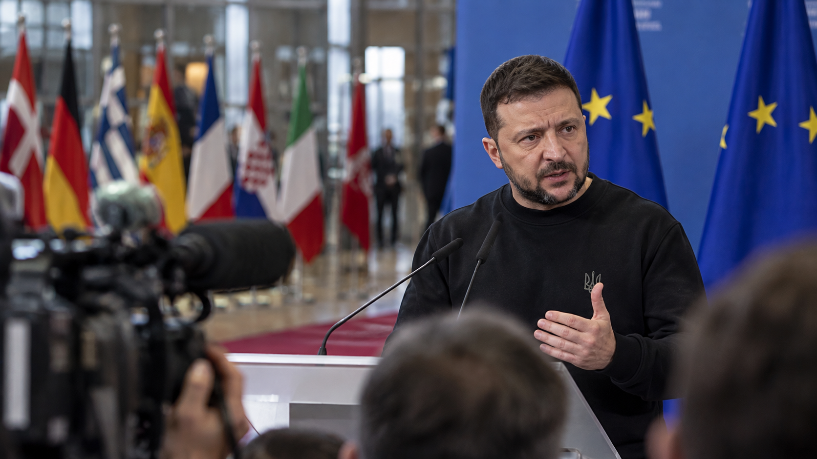 Ukrainian President Volodymyr Zelenskyy speaking at a press conference at an EU summit in Brussels, with European and EU flags behind him.